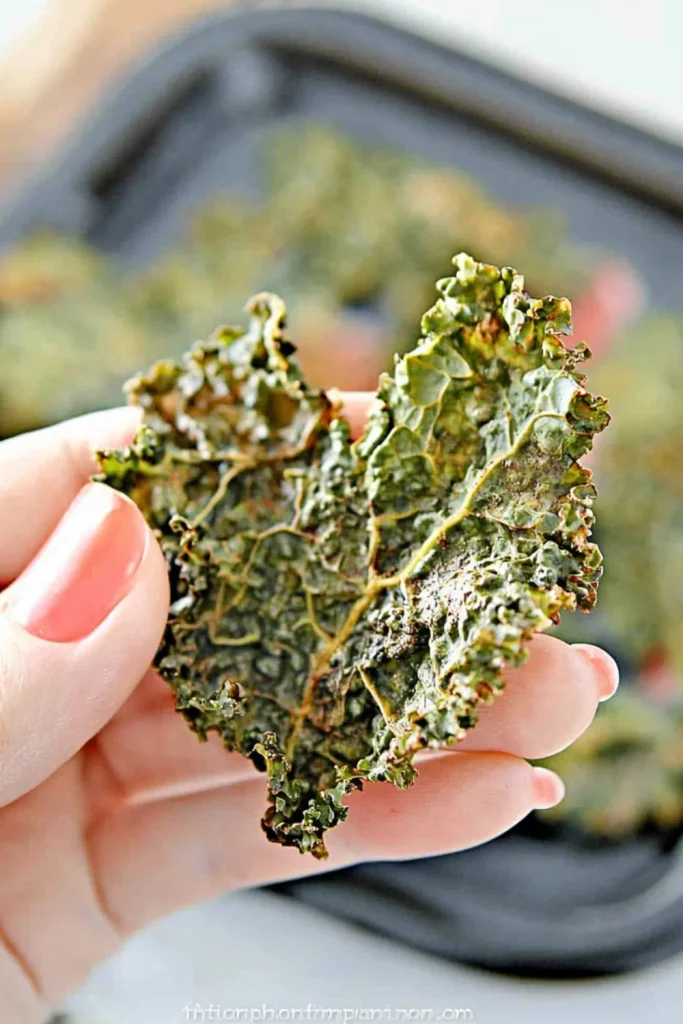 Nahaufnahme einer knusprigen, gerösteten Grünkohlchip in der Hand einer Person mit rosa Nagellack, im Hintergrund ein Backblech mit weiteren Chips.